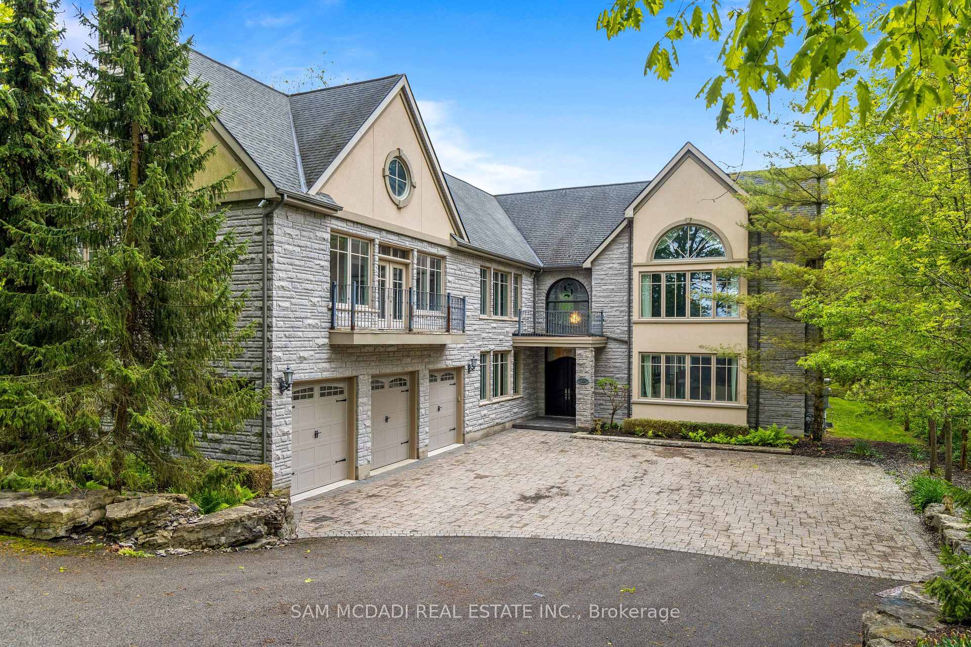 Large stone-and-stucco house with three-car garage and paved driveway among mature trees.
