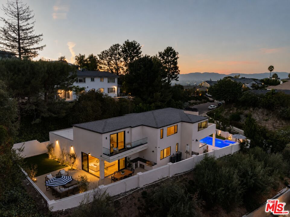 Modern two-story home with illuminated windows, a blue pool, and mountain views at dusk.