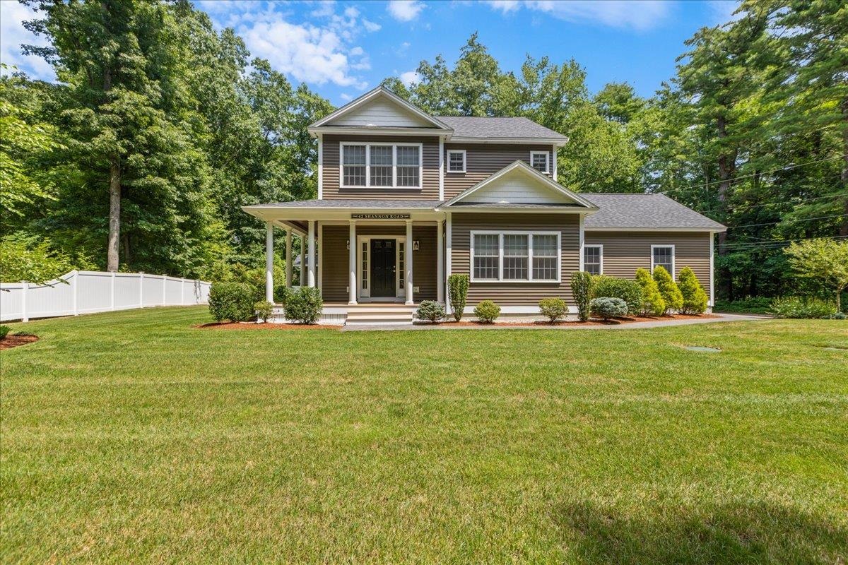 Two-story brown-sided home with a covered porch, landscaped shrubs, broad lawn, and white fence.