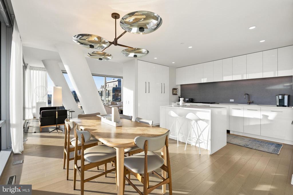 Modern white kitchen with island and bar stools beside a wooden dining table and large windows.