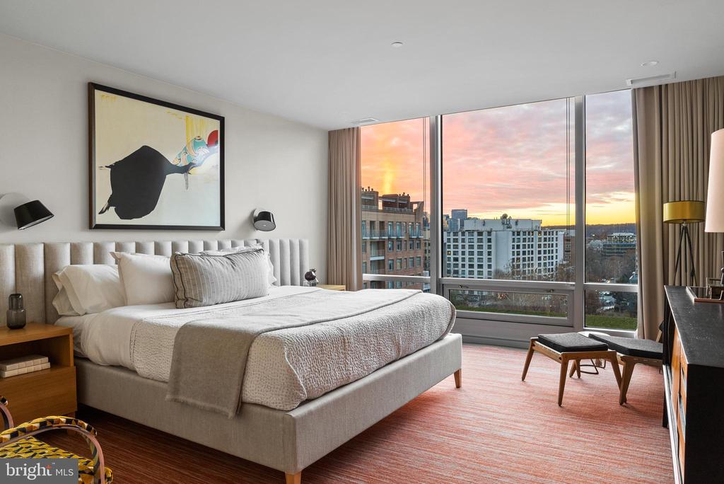 Bedroom with a king-size bed and floor-to-ceiling windows overlooking city buildings at sunset.
