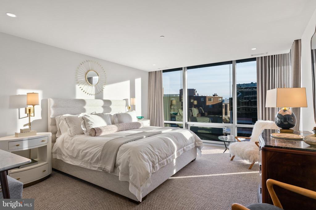 Furnished bedroom with a large upholstered bed and expansive windows overlooking nearby city buildings.