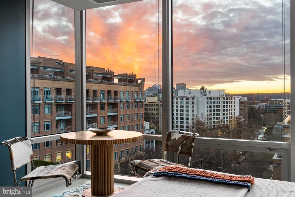 Bedroom with corner windows overlooking city buildings beneath a vivid sunset.