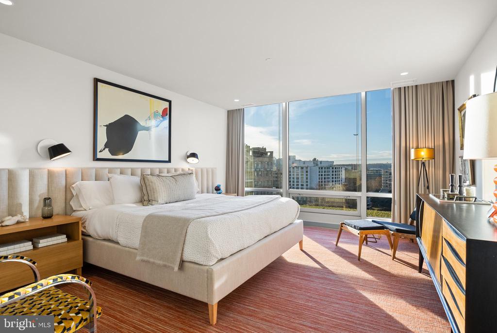 Furnished bedroom with a king-size bed, colorful artwork, and floor-to-ceiling windows overlooking nearby buildings.
