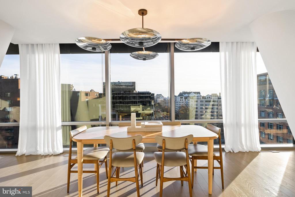 Dining area with a wood table, sculptural pendant light, and floor-to-ceiling windows overlooking city buildings.