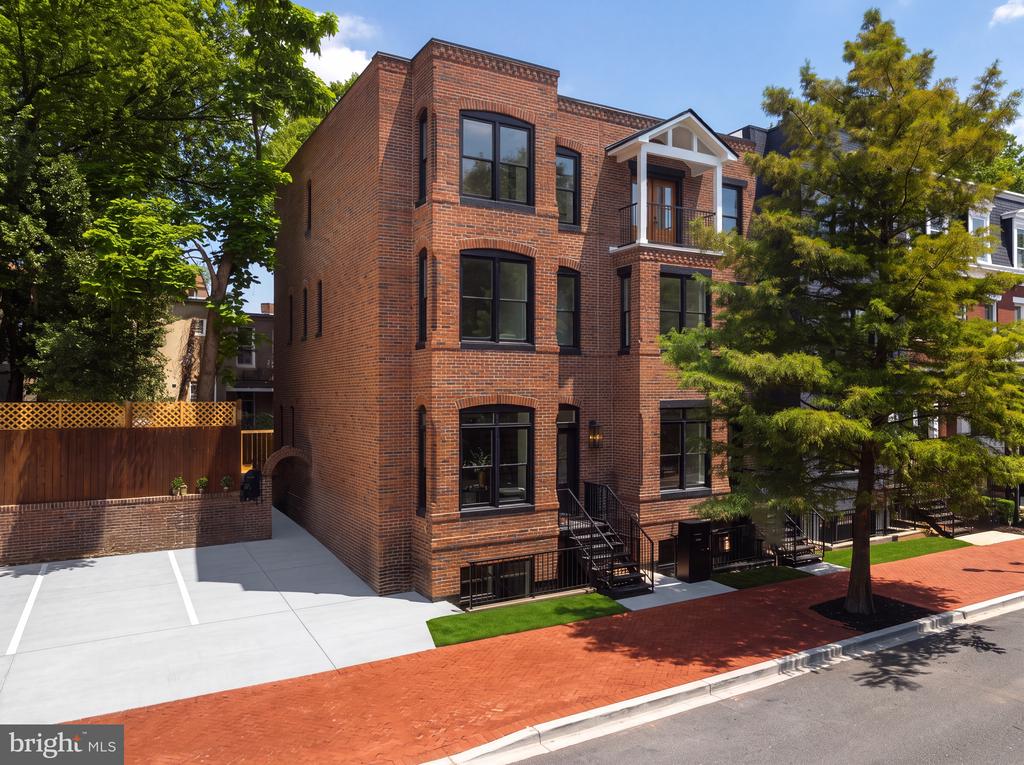 Red-brick residential building with a white balcony canopy and paved parking area beneath leafy trees.