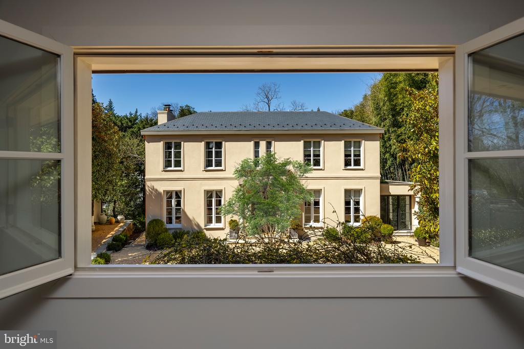 Two-story cream house with a dark roof, seen through an open window amid trees and landscaping.