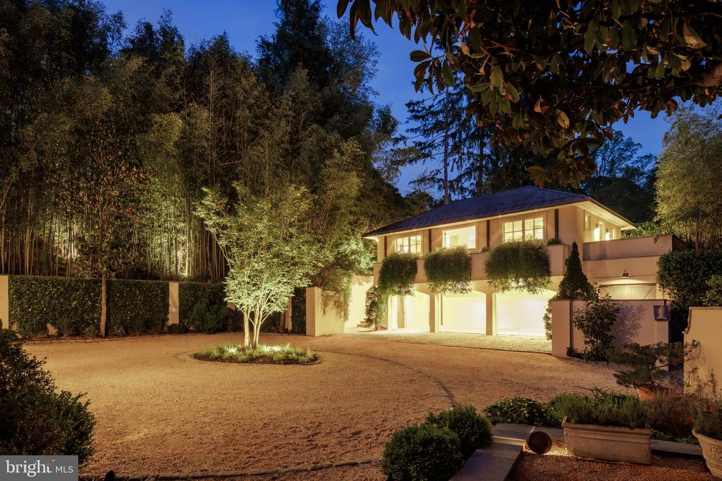 Modern two-story home lit at dusk, framed by mature trees and a broad gravel driveway.