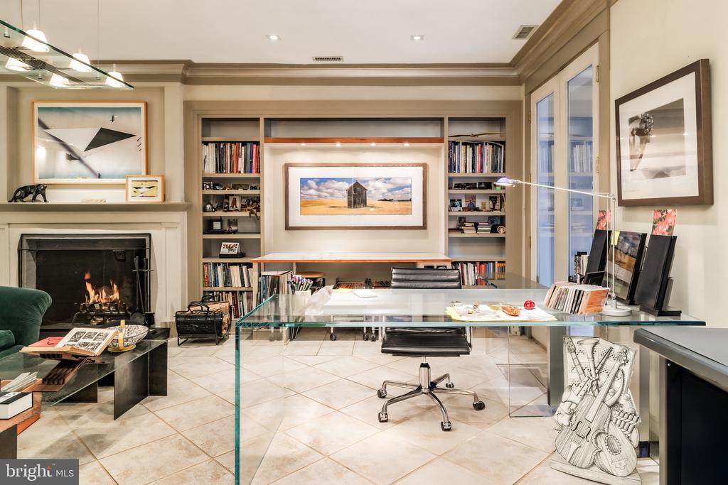 Home office with built-in bookshelves, a glass desk, and a lit fireplace.