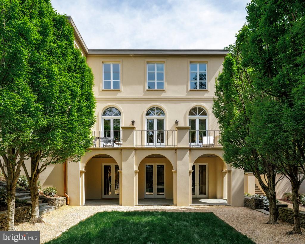 Rear of a cream-colored home with arched openings, a balcony, and tall trees framing the lawn.