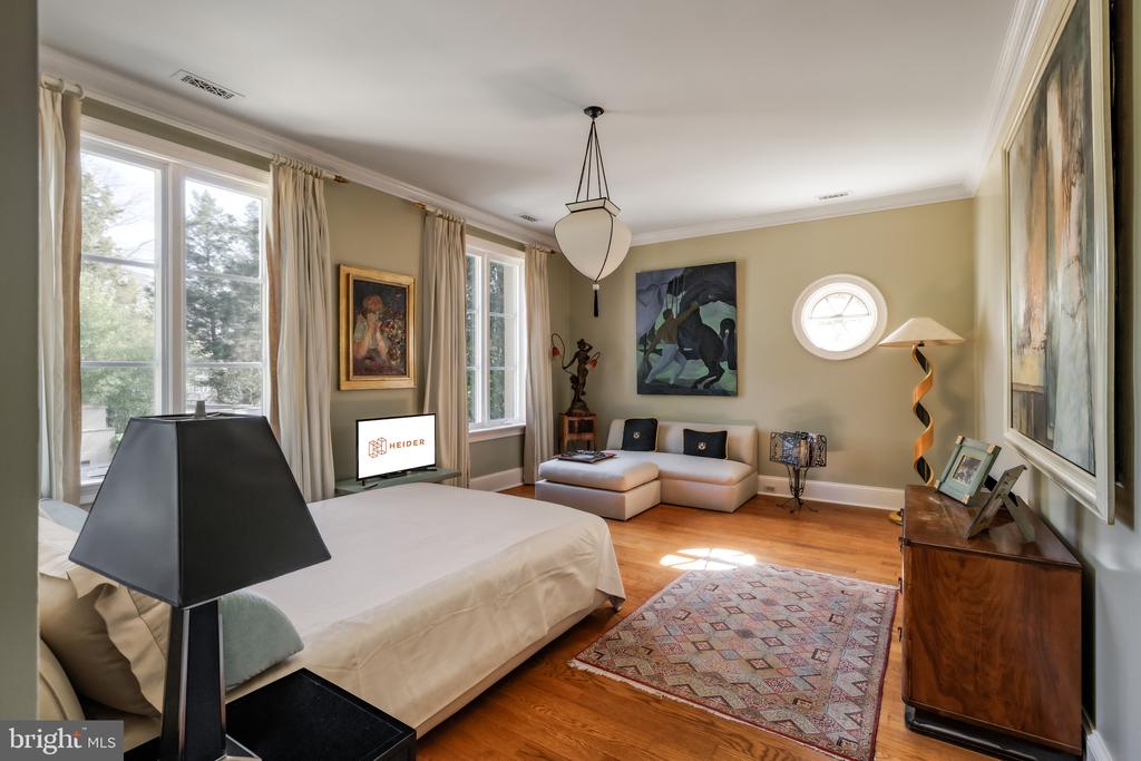 Bedroom with a white bed, tall curtained windows, cream sectional sofa, hardwood floors, and patterned rug.