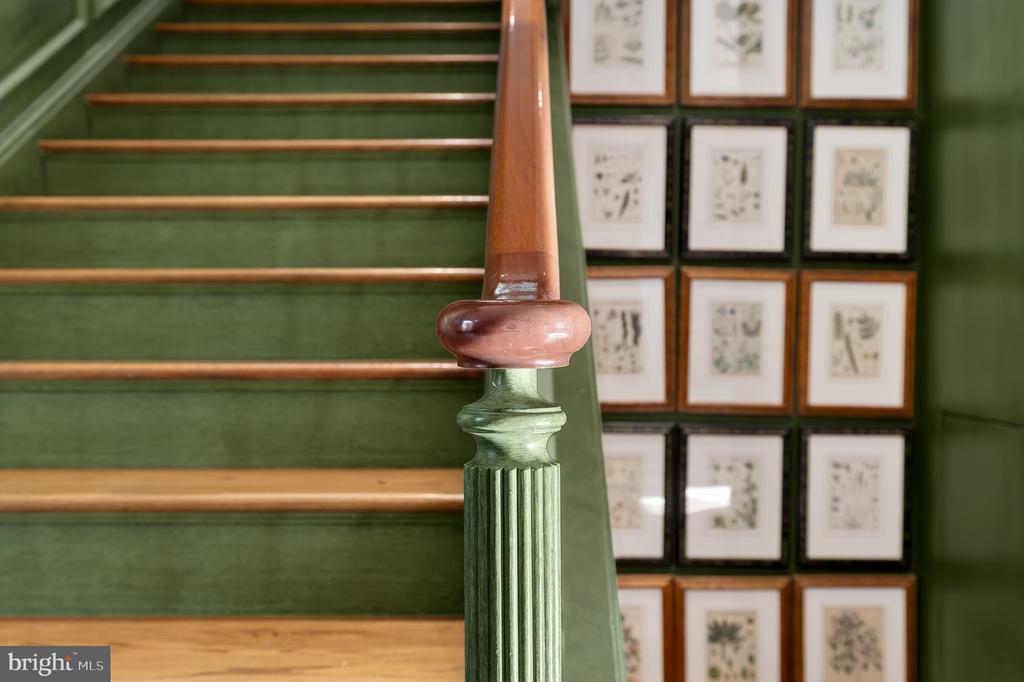 Green carpeted staircase with wood treads, a fluted newel post, and framed botanical prints.