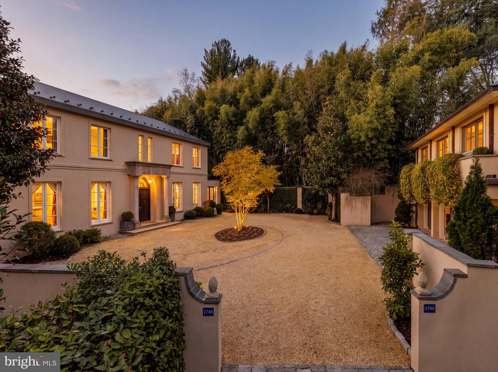 Two-story residence with glowing windows around a gravel courtyard and ornamental tree at dusk.