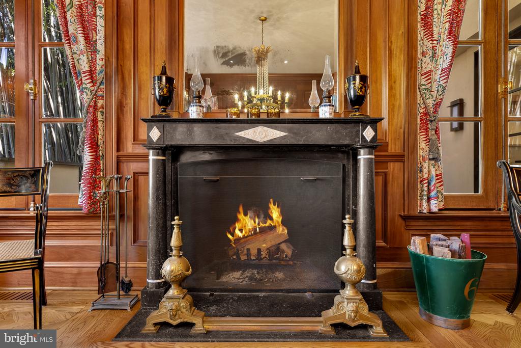 Ornate marble fireplace with a lit fire, mirrored mantel, wood paneling, and patterned curtains.