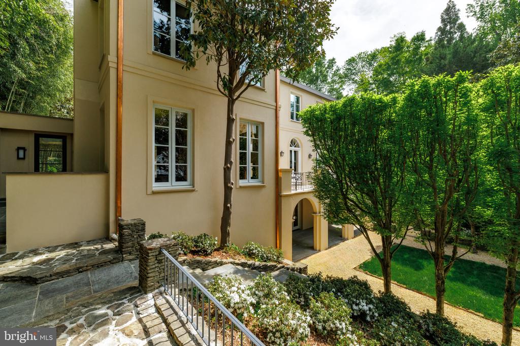 Beige home with a stone terrace, arched driveway entrance, and mature trees.