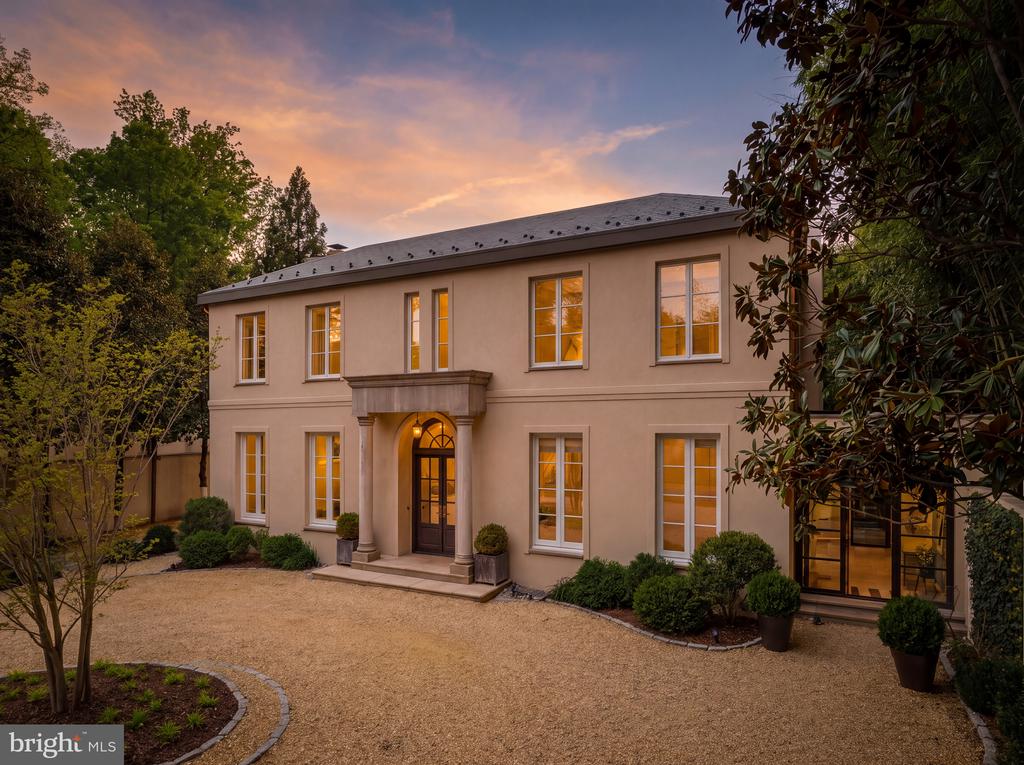 Two-story beige home with a columned entry and warmly lit windows at dusk.