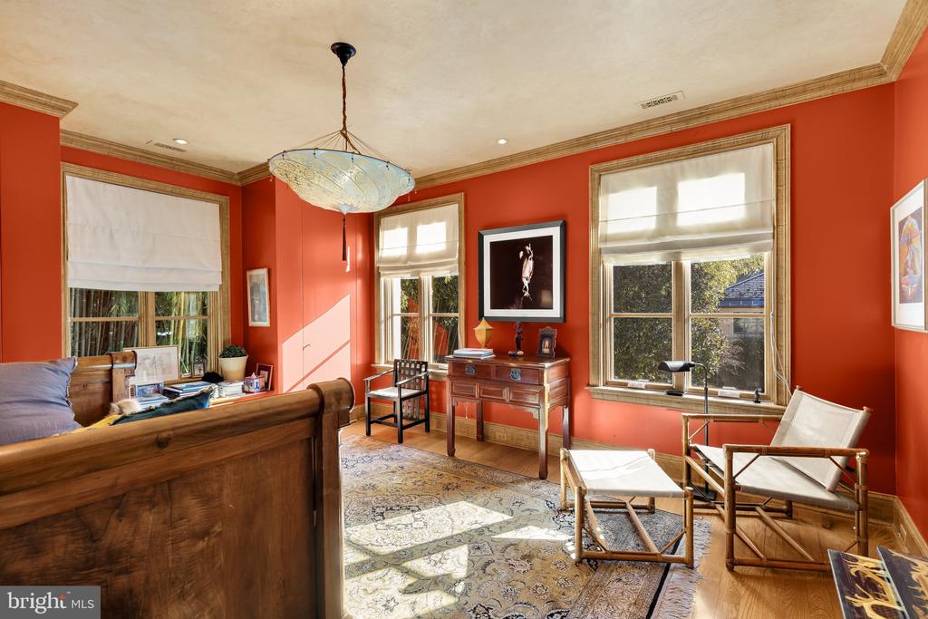 Sunlit sitting room with coral-red walls, tall windows, a wood desk, and a patterned rug.