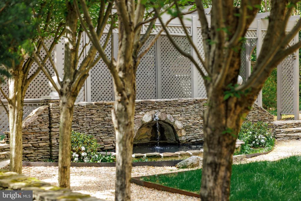 Garden fountain flowing into a pond behind a low stone wall and white lattice fence.