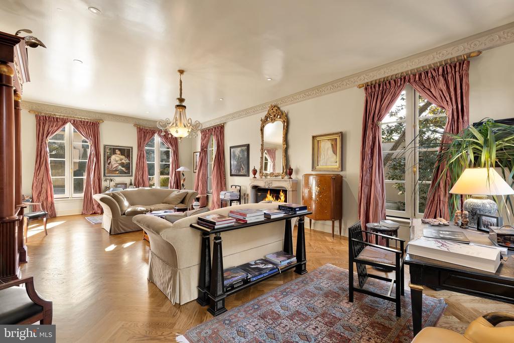 Formal living room with parquet floors, fireplace, chandelier, and tall windows with rose-colored curtains.