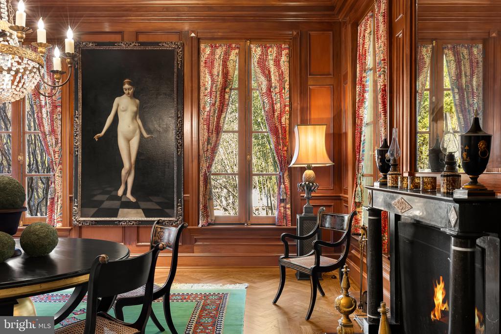 Wood-paneled dining room with a marble fireplace, tall curtained windows, and a round table.