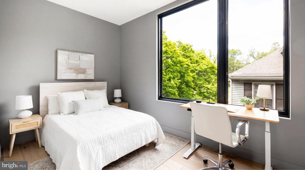 Bedroom with a white bed and desk beside a large window overlooking leafy trees.