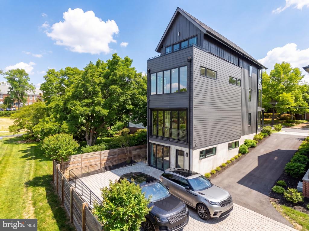 Modern gray three-story house with a steep roof, fenced patio, and two parked vehicles.