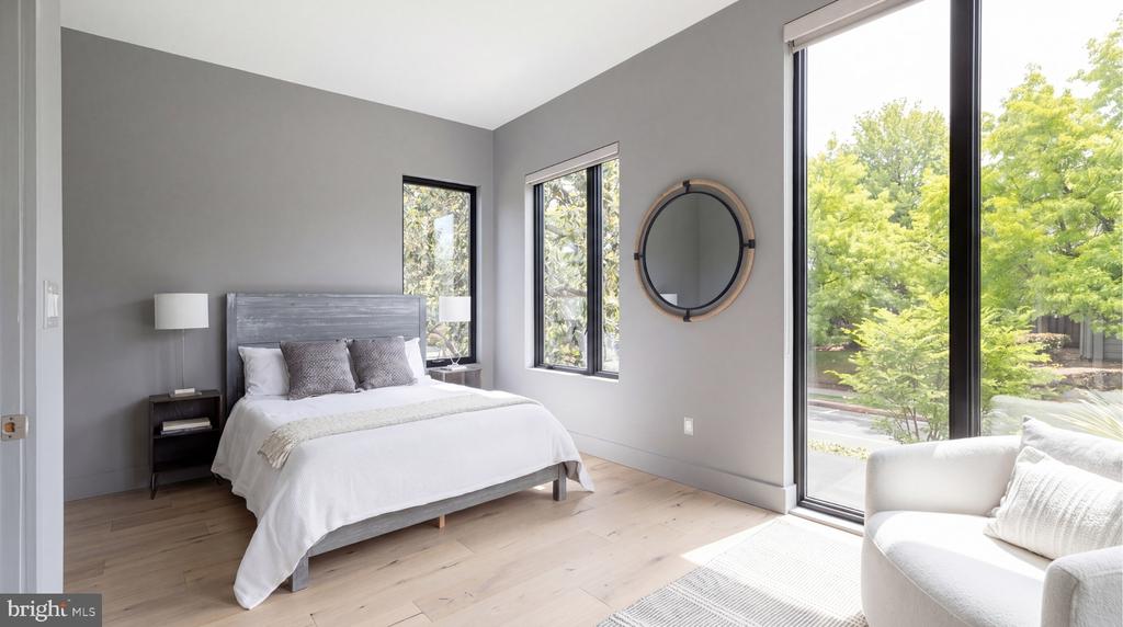 Bright bedroom with a gray bed, light wood floors, and large windows overlooking trees.