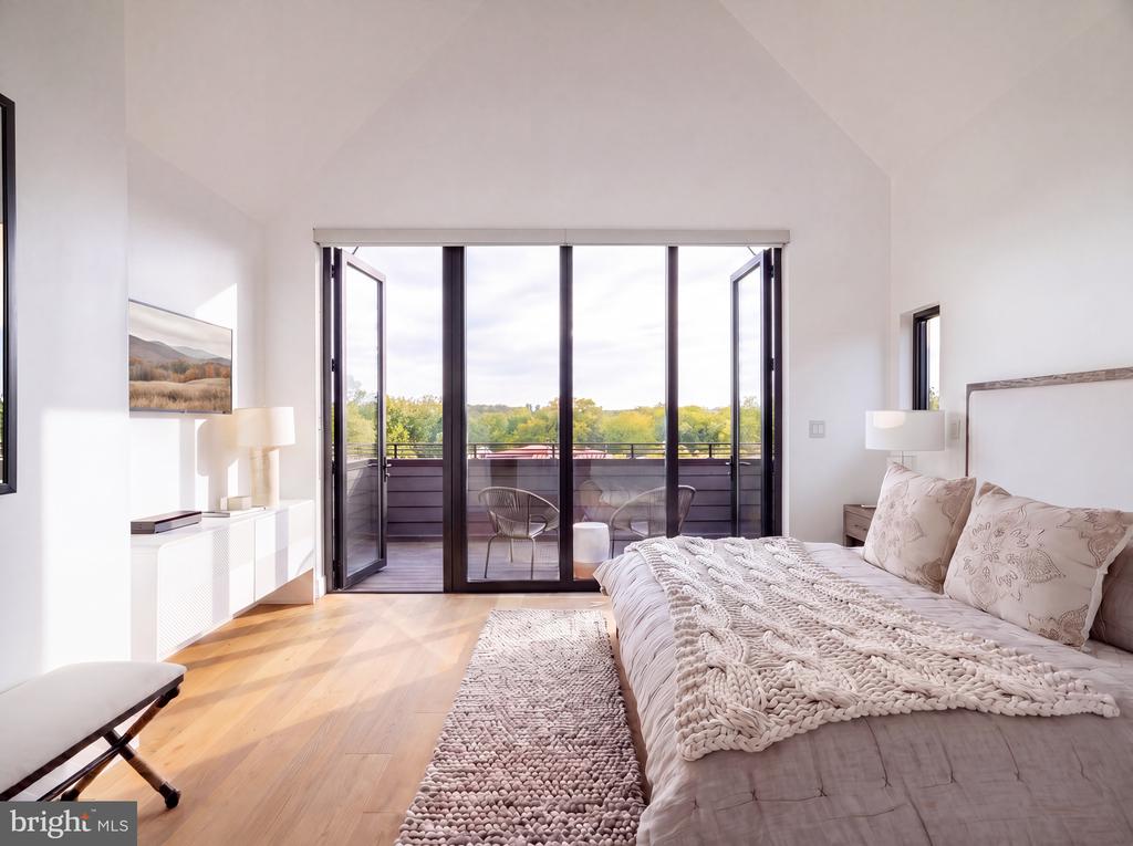 Furnished bedroom with tall glass doors opening onto a balcony overlooking trees.