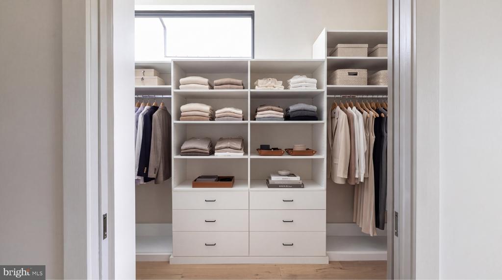 Walk-in closet with white built-ins, neatly arranged clothes, and a high window.
