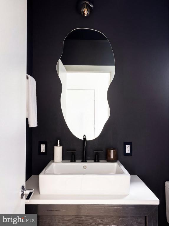 Bathroom vanity with a white vessel sink, black fixtures, and an irregularly shaped mirror against a dark wall.