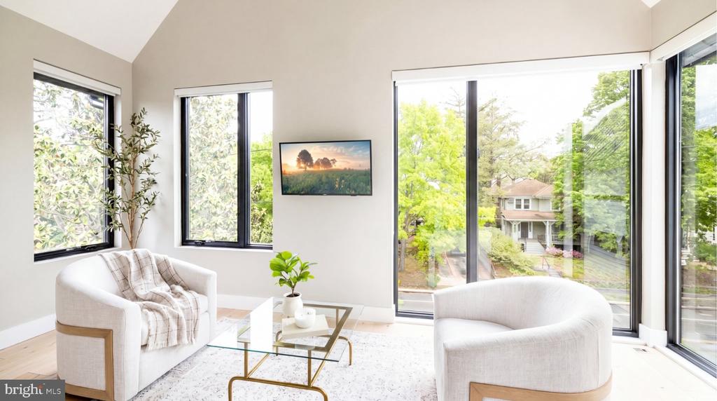 Bright sitting area with white chairs, a glass coffee table, and large windows overlooking trees.