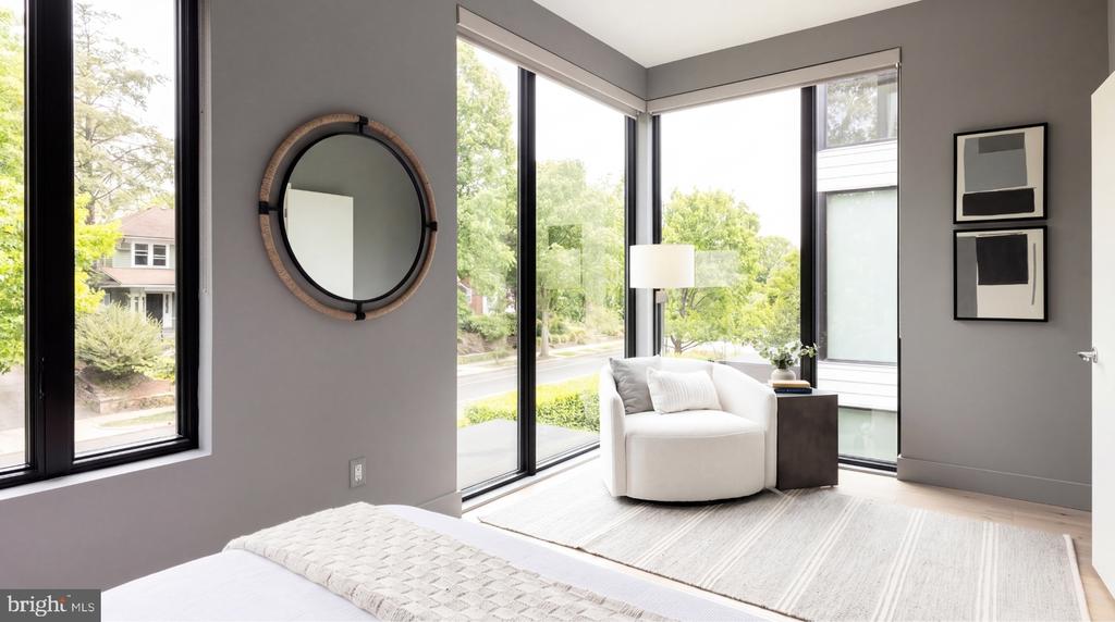 Staged bedroom with a white bed, rounded armchair, and tall corner windows overlooking trees.
