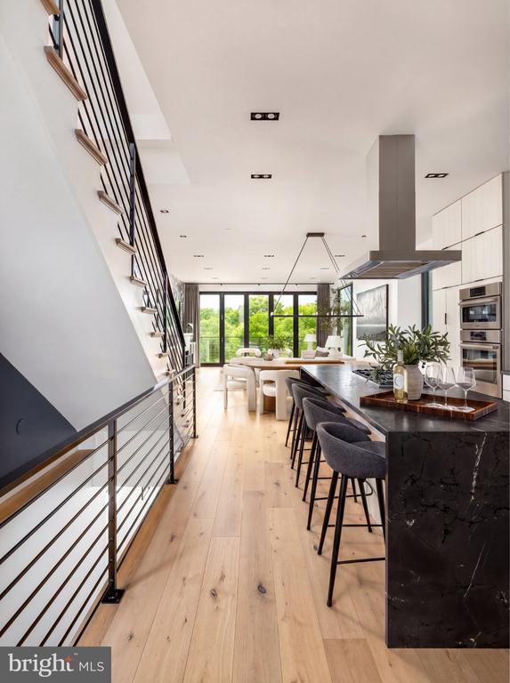 Modern kitchen with a dark stone island, black-railed staircase, and tall windows overlooking trees.