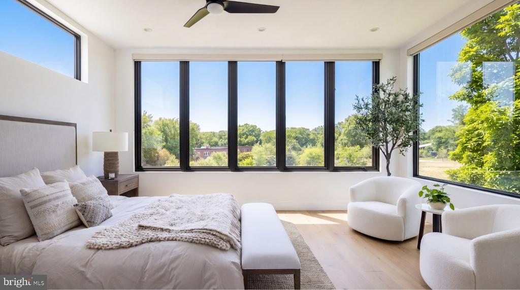 Bright bedroom with a large bed, white chairs, and broad windows overlooking leafy greenery.