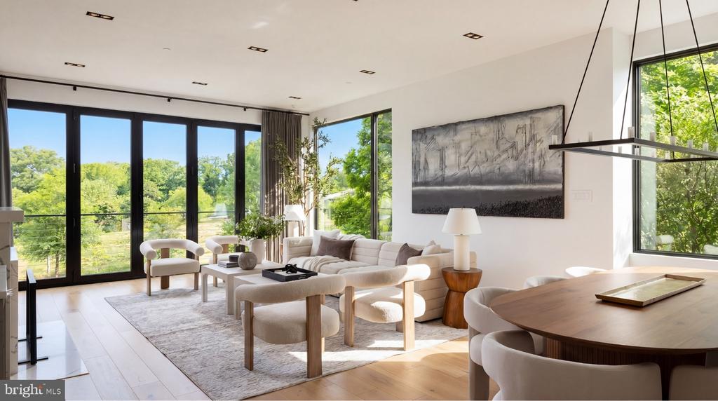 Modern living and dining room with floor-to-ceiling windows overlooking green trees.