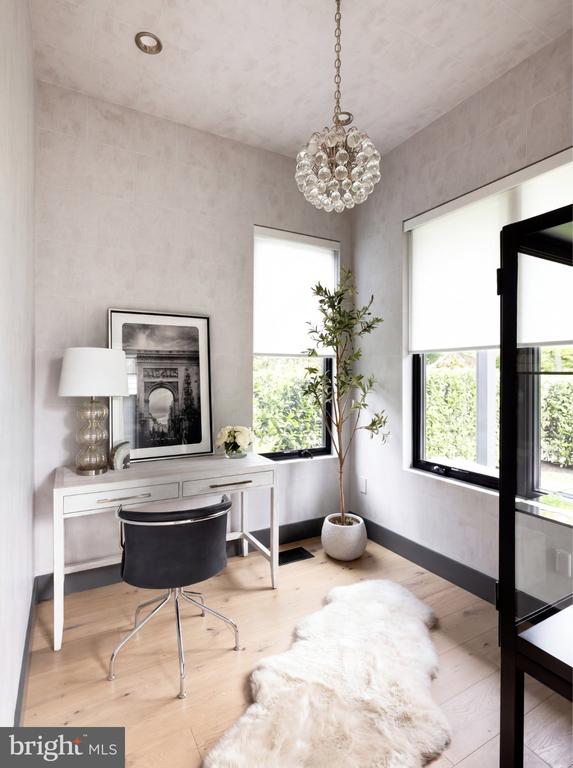 Furnished home office with a white desk, black chair, crystal pendant, and tall windows.