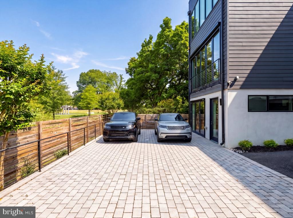 Modern home beside a broad paver driveway with two parked vehicles and a wood-and-metal fence.