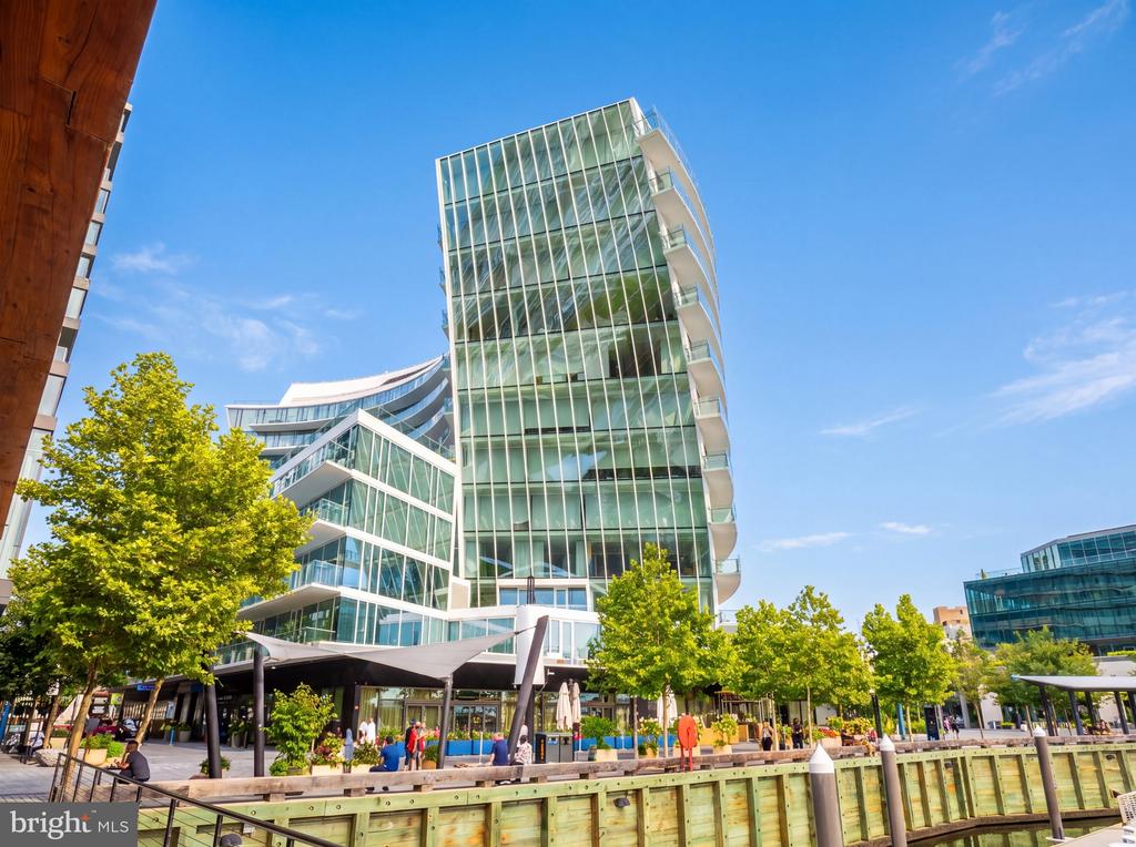 Modern glass condominium tower beside a tree-lined waterfront promenade.