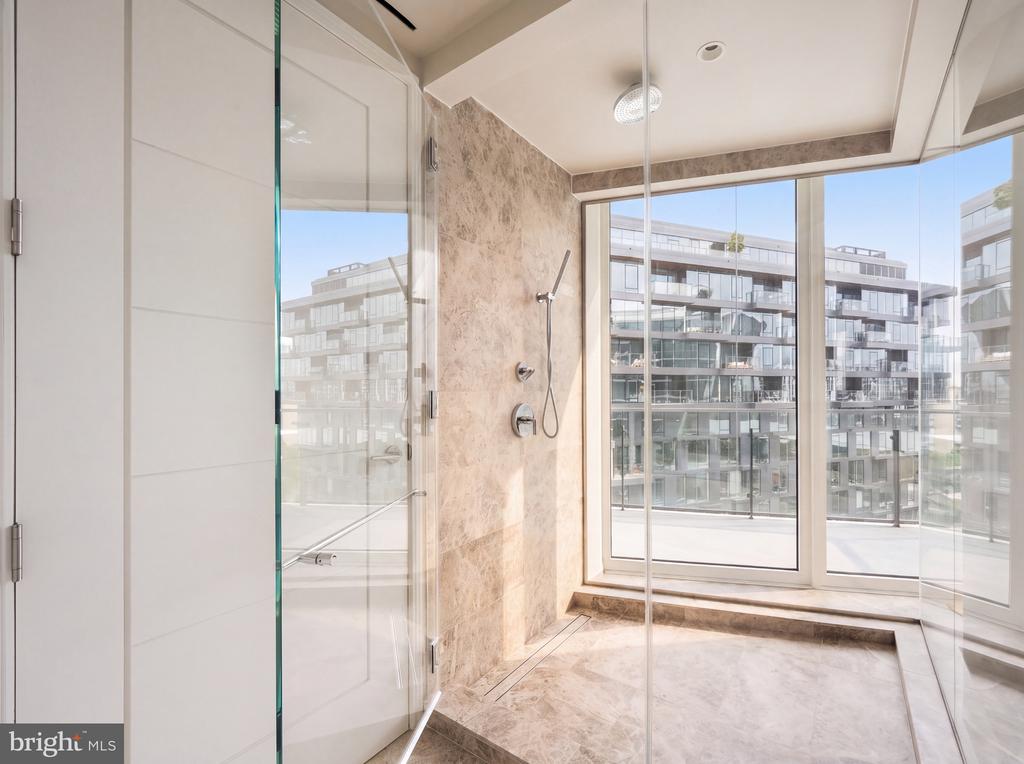 Marble walk-in shower with glass walls and large windows overlooking modern apartment buildings.