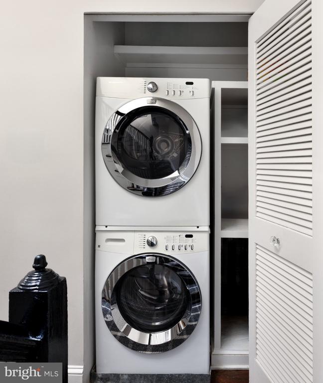 Laundry closet with stacked white front-loading washer and dryer beside open shelving.