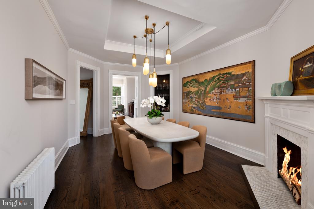 Dining room with a long white table, tan chairs, landscape painting, and lit fireplace.