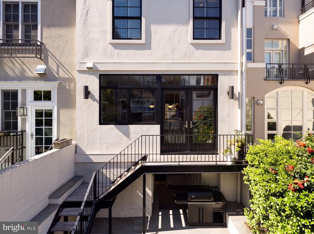 Townhouse rear courtyard with black metal stairs, a raised landing, outdoor grill, and flowering shrubs.