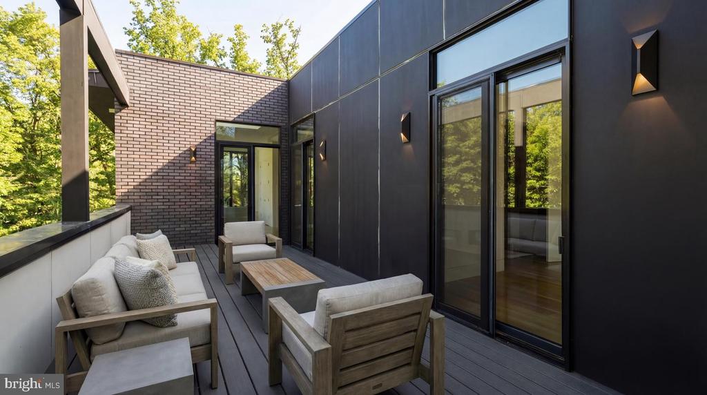 Modern home terrace with wood lounge furniture, gray decking, brick wall, and large glass doors.