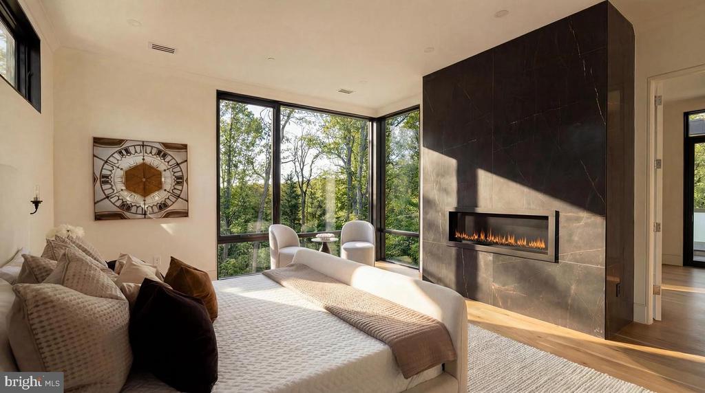 Bedroom with a dark stone fireplace, large bed, and corner windows overlooking trees.