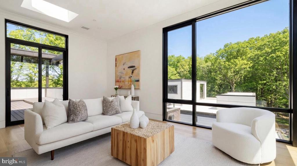 Staged living room with a white sofa, skylight, and large windows overlooking leafy trees.