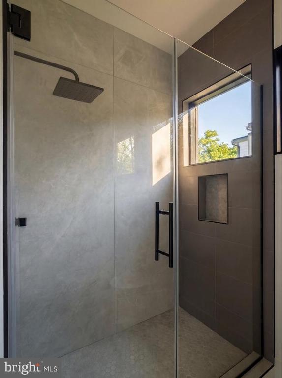 Walk-in shower with a glass enclosure, dark rainfall showerhead, tiled walls, built-in niche, and high window.