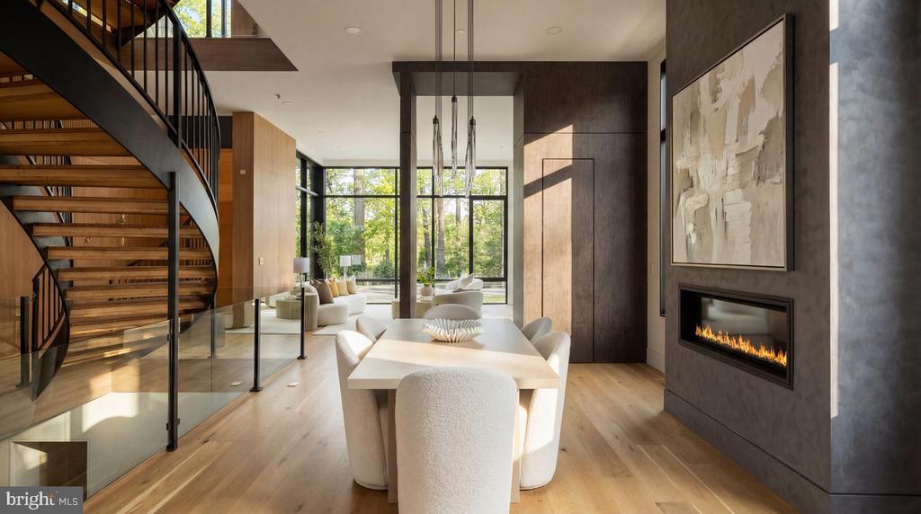 Modern dining room with a sculptural staircase, linear fireplace, and windows overlooking greenery.