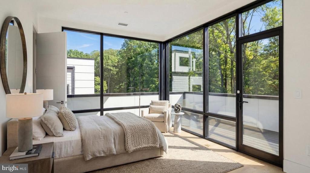 Modern bedroom with a neutral bed and floor-to-ceiling windows overlooking leafy trees.