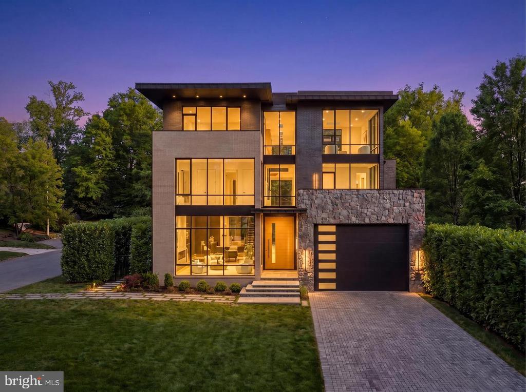 Modern two-story home at dusk with glowing windows, stone garage facade, and paved driveway.