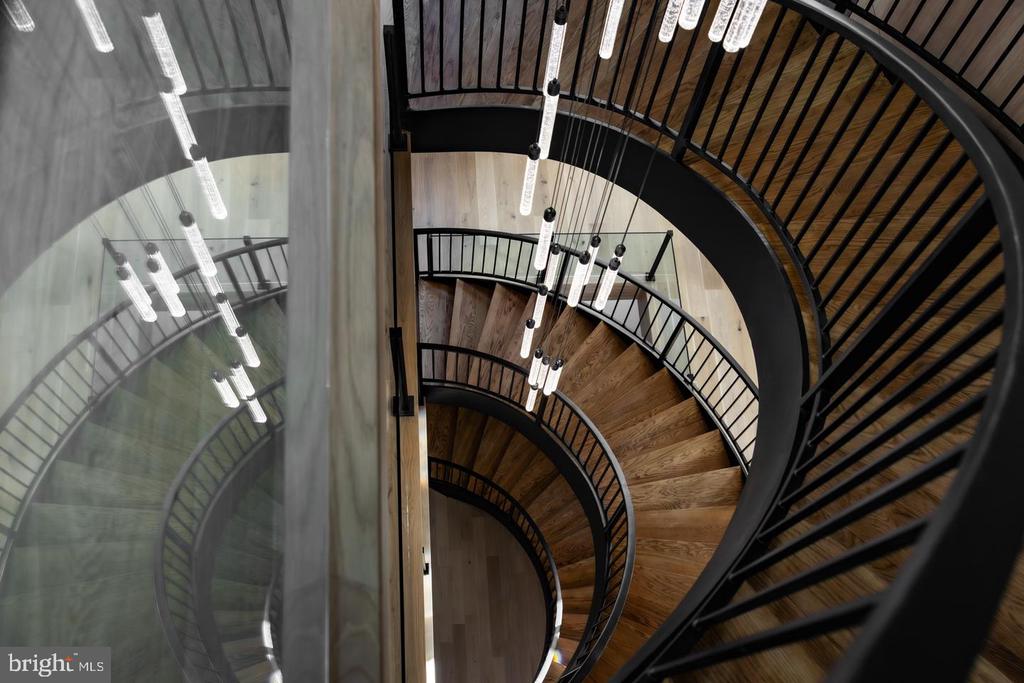 Curved wood staircase with black railings and suspended lights, viewed from above.